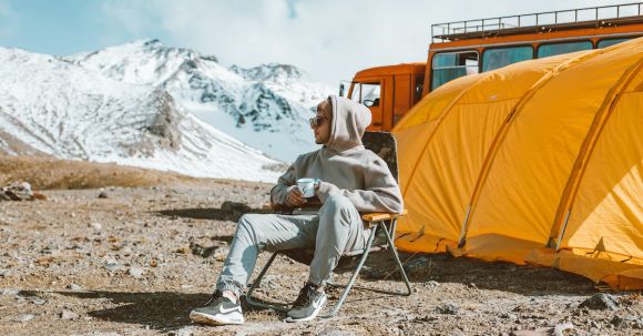 Tranquil Getaways - Full length of male camper sitting on folding chair next to tent in middle of mountain valley and enjoying views