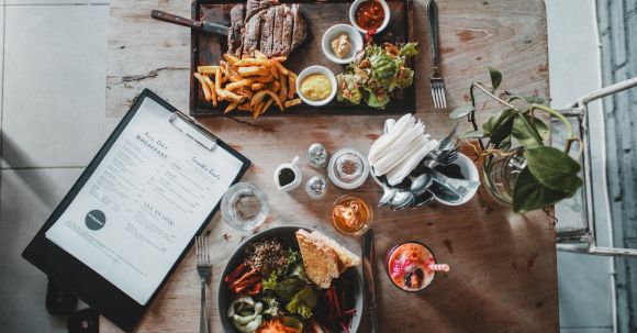 French Culinary Journey - Top view of wooden table with salad bowl and fresh drink arranged with tray of appetizing steak and french fries near menu in cozy cafe