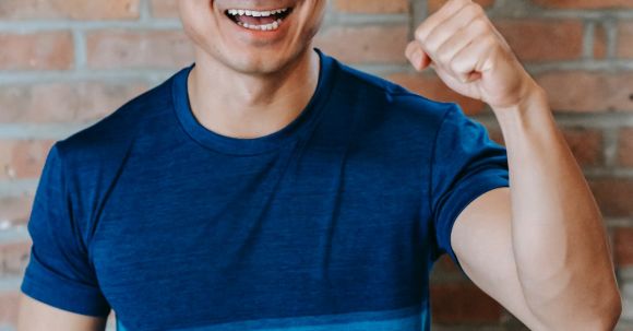 Fitness Success - Cheerful young ethnic male athlete in sportswear smiling and showing fist while standing against brick wall after successful workout in gym