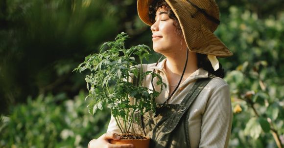 Mindfulness Cultivation - Content ethnic female grower in panama hat with potted plant and closed eyes in summer garden