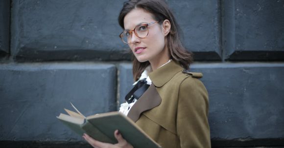 Develop Style - From below confident young female in glasses and elegant vintage clothes looking away and contemplating while reading rook on street near black wall of aged building in city