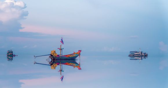 Harmony In Fishing - Traditional colorful fishing boat floating on surface of calm sea near modern vessels against sunset sky reflecting in blue water